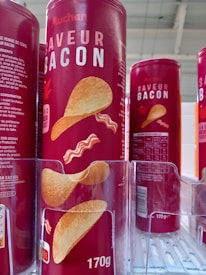 Cylindrical red containers with bold white text displaying 'SAVEUR BACON' and an image of chips and bacon strips are lined up on a shelf. The packaging indicates a 170g size with nutritional information and branding visible.