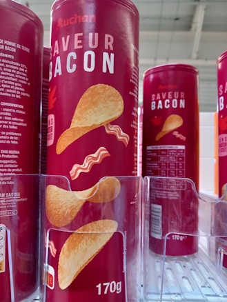 Cylindrical red containers with bold white text displaying 'SAVEUR BACON' and an image of chips and bacon strips are lined up on a shelf. The packaging indicates a 170g size with nutritional information and branding visible.