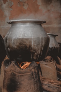 A large metal pot is sitting on a traditional clay stove with an open flame underneath, emitting steam. The background features a textured, worn wall, adding to the rustic atmosphere.