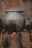A large metal pot is sitting on a traditional clay stove with an open flame underneath, emitting steam. The background features a textured, worn wall, adding to the rustic atmosphere.