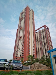 A tall residential high-rise building with red and beige colors stands prominently against a partly cloudy sky. In the foreground, several cars are parked next to a small landscaped area with greenery. A pile of bricks is also visible, suggesting ongoing construction or landscaping work nearby.