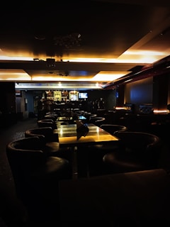 A dimly lit bar area with several tables and leather chairs arranged in rows. The ceiling features recessed lighting that casts a warm glow over the scene. The bar itself is in the background, with bottles of alcohol displayed on shelves and a bartender standing behind the counter. There is a television mounted on the wall behind the bar.