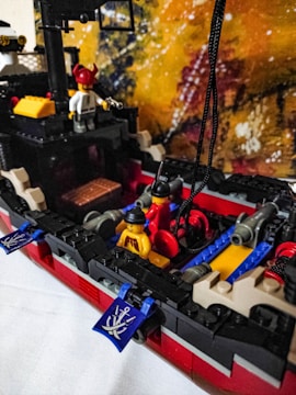 A detailed LEGO pirate ship scene with mini-figures, featuring a deck with cannons, a mast, and a colorful background. The mini-figures are dressed in pirate-themed attire including hats and torsos decorated with pirate insignia. The ship has red and black sections, with blue flags displaying crossbones.