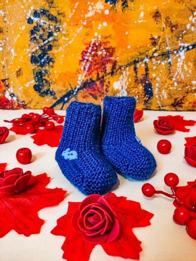 A pair of knitted blue baby booties is placed on a surface adorned with red artificial roses and red berries. In the background, an abstract painting with vibrant yellow, orange, and dark blue hues adds visual contrast.