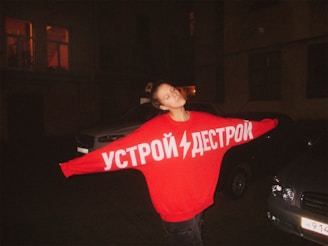 A person stands outdoors at night, wearing an oversized red sweater with white Cyrillic text on it. The person has their arms outstretched and eyes closed, appearing relaxed or content. Surrounding them are parked cars, and the dimly lit background includes building windows with some lights on.