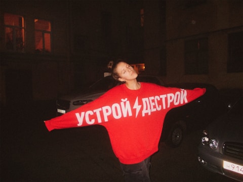 A person stands outdoors at night, wearing an oversized red sweater with white Cyrillic text on it. The person has their arms outstretched and eyes closed, appearing relaxed or content. Surrounding them are parked cars, and the dimly lit background includes building windows with some lights on.