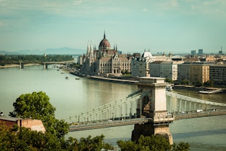 A picturesque view of a historic European cityscape featuring a prominent river and bridge. The parliament building with its distinctive Gothic architecture stands out prominently along the riverbank. Trees and urban buildings line the horizon under a serene sky.