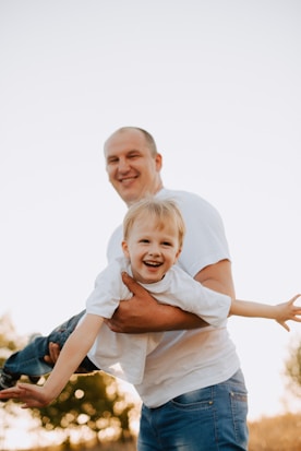 A man, dressed in a white T-shirt and blue jeans, is holding a smiling young boy, also in a white T-shirt, who appears to be flying or playing airplane. The background shows a natural outdoor setting with soft sunlight and some blurred trees.