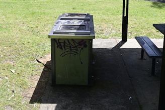 A public barbecue grill with a graffiti design on its side, situated on a concrete surface next to a grassy area. Part of a picnic table is visible in the right corner. Sunlight and shadows play across the scene.