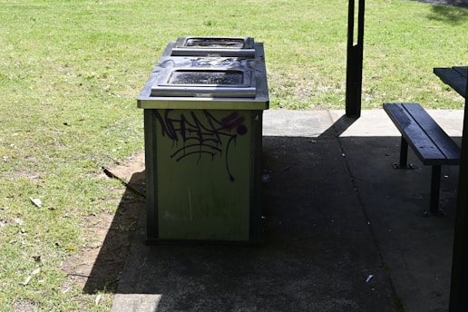 A public barbecue grill with a graffiti design on its side, situated on a concrete surface next to a grassy area. Part of a picnic table is visible in the right corner. Sunlight and shadows play across the scene.