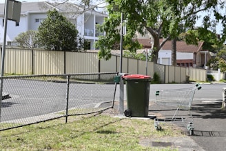 A suburban street scene featuring a green trash bin with a red lid and an empty shopping cart standing on an asphalt surface. A chain-link fence separates the sidewalk from a grassy area. Tree branches and leaves provide some shade, while several residential houses are visible in the background, bordered by light-colored fences and walls. Overhead, the sky is clear with a few traces of blue.