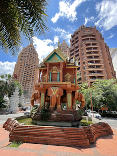 A decorative pavilion with intricate design stands prominently, featuring multiple carved elephants at its base. The structure is set against a backdrop of tall, modern buildings with reddish-brown exteriors. The sky is clear with scattered clouds, and lush greenery surrounds the area, enhancing the vibrant urban setting.