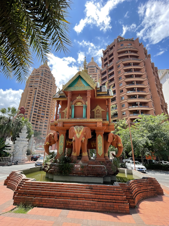 A decorative pavilion with intricate design stands prominently, featuring multiple carved elephants at its base. The structure is set against a backdrop of tall, modern buildings with reddish-brown exteriors. The sky is clear with scattered clouds, and lush greenery surrounds the area, enhancing the vibrant urban setting.
