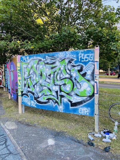 A vibrant graffiti mural featuring abstract designs in blue, green, and white colors displayed on a wooden panel. The artwork is set against a backdrop of leafy trees and an adjacent street. Spray paint cans and other materials are scattered on the ground, suggesting recent activity.