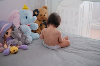 A young child wearing a diaper is sitting on a bed with their back facing the camera. Surrounding the child are several plush toys, including a large blue elephant, a brown teddy bear, a doll wearing a purple hat, and a small gray elephant. The setting appears to be a cozy bedroom with soft lighting and a crib visible in the background.