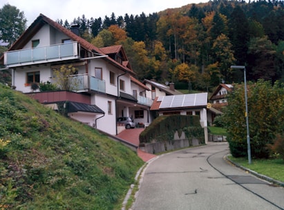 A residential house with white walls and brown roofs, set against a hillside covered with autumn foliage. The house has multiple levels with balconies and solar panels on the roof. There's a winding road leading up to the house, and the surrounding area is lush with trees and greenery.