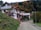 A residential house with white walls and brown roofs, set against a hillside covered with autumn foliage. The house has multiple levels with balconies and solar panels on the roof. There's a winding road leading up to the house, and the surrounding area is lush with trees and greenery.
