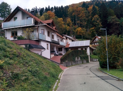 A residential house with white walls and brown roofs, set against a hillside covered with autumn foliage. The house has multiple levels with balconies and solar panels on the roof. There's a winding road leading up to the house, and the surrounding area is lush with trees and greenery.