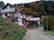 A residential house with white walls and brown roofs, set against a hillside covered with autumn foliage. The house has multiple levels with balconies and solar panels on the roof. There's a winding road leading up to the house, and the surrounding area is lush with trees and greenery.