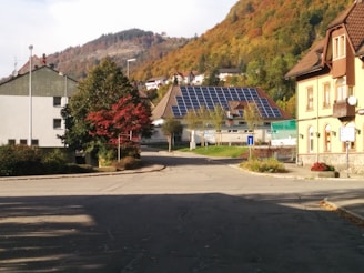 A quiet residential street surrounded by houses with colorful autumn foliage on the trees. Solar panels are installed on the roof of one of the houses in the background. The area is calm and well-maintained.