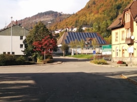 A quiet residential street surrounded by houses with colorful autumn foliage on the trees. Solar panels are installed on the roof of one of the houses in the background. The area is calm and well-maintained.