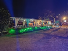 An outdoor event space is decorated with draped white fabric, tables, and chairs under a canopy. Green lighting illuminates the surrounding greenery, creating a vibrant atmosphere. Several people are visible in the background, suggesting a social gathering.