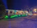An outdoor event space is decorated with draped white fabric, tables, and chairs under a canopy. Green lighting illuminates the surrounding greenery, creating a vibrant atmosphere. Several people are visible in the background, suggesting a social gathering.