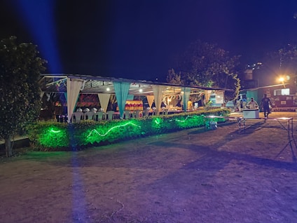 An outdoor event space is decorated with draped white fabric, tables, and chairs under a canopy. Green lighting illuminates the surrounding greenery, creating a vibrant atmosphere. Several people are visible in the background, suggesting a social gathering.