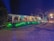 An outdoor event space is decorated with draped white fabric, tables, and chairs under a canopy. Green lighting illuminates the surrounding greenery, creating a vibrant atmosphere. Several people are visible in the background, suggesting a social gathering.
