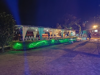 An outdoor event space is decorated with draped white fabric, tables, and chairs under a canopy. Green lighting illuminates the surrounding greenery, creating a vibrant atmosphere. Several people are visible in the background, suggesting a social gathering.