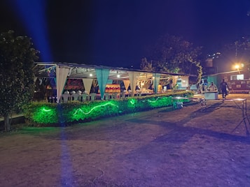 An outdoor event space is decorated with draped white fabric, tables, and chairs under a canopy. Green lighting illuminates the surrounding greenery, creating a vibrant atmosphere. Several people are visible in the background, suggesting a social gathering.