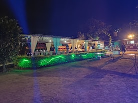 An outdoor event space is decorated with draped white fabric, tables, and chairs under a canopy. Green lighting illuminates the surrounding greenery, creating a vibrant atmosphere. Several people are visible in the background, suggesting a social gathering.