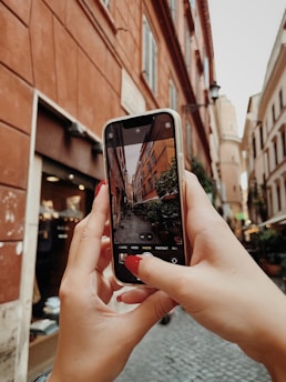 The photo captures a close-up of hands holding a smartphone, which is focused on capturing an image of a narrow street lined with tall, historic buildings. The scene on the phone screen mirrors the buildings and cobblestone path seen in the background. The surrounding architecture features warm, earthy tones, and there's a sense of depth created by the receding buildings.