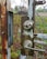 A close-up of a rusted metal gate with a worn-out lock mechanism. The lock and handle are attached to a chain-link fence, with visible peeling paint and moss growth. Behind the gate, there's overgrown vegetation and a field in the distance, showcasing a natural setting.