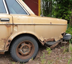 A rusted and damaged car with peeling paint, particularly around the wheel arch. The tire appears worn, and the overall condition suggests the vehicle has been neglected for an extended period. Surrounding the car, there is greenery, indicating it is parked outdoors near vegetation.