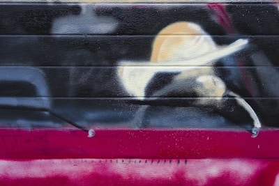 Graffiti art depicting a minimalist portrayal of a person wearing a wide-brimmed hat. The image is spray-painted on a horizontal metal surface, blending stark contrasts of light and dark colors with an abstract style.