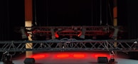 A dimly lit stage with suspended lighting equipment featuring multiple spotlights and cables. The floor is illuminated by red lights, creating a dramatic atmosphere.