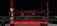 A dimly lit stage with suspended lighting equipment featuring multiple spotlights and cables. The floor is illuminated by red lights, creating a dramatic atmosphere.