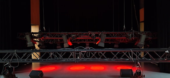 A dimly lit stage with suspended lighting equipment featuring multiple spotlights and cables. The floor is illuminated by red lights, creating a dramatic atmosphere.