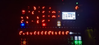 A dark environment illuminated by numerous glowing buttons and knobs on an electronic device, featuring a mix of red, blue, green, and white lights. There's a small screen displaying various rectangular labels.
