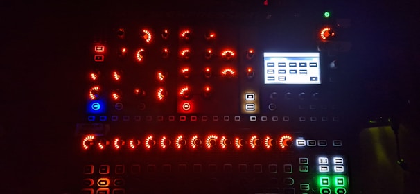 A dark environment illuminated by numerous glowing buttons and knobs on an electronic device, featuring a mix of red, blue, green, and white lights. There's a small screen displaying various rectangular labels.