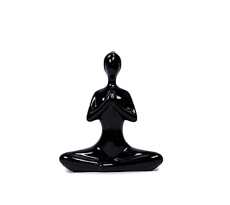 A sleek, black sculpture of a human figure seated in a meditative pose with hands in a prayer position. The figure is minimalist, with smooth surfaces and an abstract design, emphasizing calmness and balance.