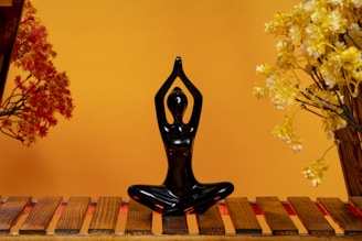 A black figurine in a yoga pose sits on a wooden platform. Surrounding the figurine are artificial plants with red and yellow leaves, set against a warm orange background.