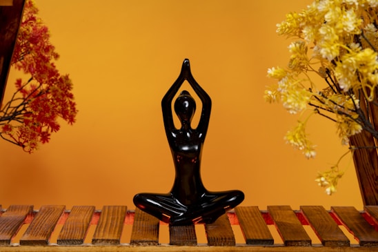 A black figurine in a yoga pose sits on a wooden platform. Surrounding the figurine are artificial plants with red and yellow leaves, set against a warm orange background.