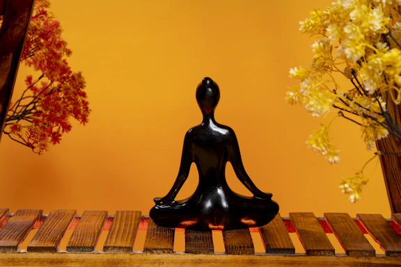 A black, glossy statue of a person sits cross-legged on a wooden surface, with branches of artificial flowers on either side. The background is a warm orange, creating a harmonious and calm atmosphere.