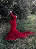 A pregnant woman in a long, flowing red dress stands in a natural setting, surrounded by green foliage and grass. Her hair is long and dark, covering part of her face as she looks around. The sunlight filters through the trees, casting a soft light on the scene.
