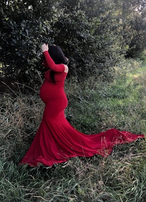 A pregnant woman in a long, flowing red dress stands in a natural setting, surrounded by green foliage and grass. Her hair is long and dark, covering part of her face as she looks around. The sunlight filters through the trees, casting a soft light on the scene.