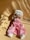 A handmade doll with long blonde hair is wearing a white knitted hat, a pink dress, and white shoes. It is laid on a beige surface with shadows cast across the scene, creating an artistic and warm ambiance.
