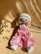 A handmade doll with long blonde hair is wearing a white knitted hat, a pink dress, and white shoes. It is laid on a beige surface with shadows cast across the scene, creating an artistic and warm ambiance.