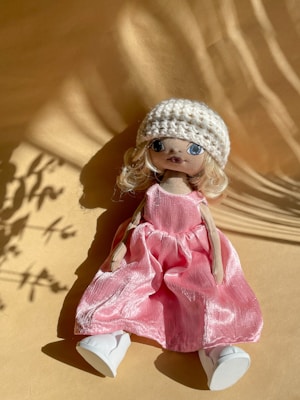 A handmade doll with long blonde hair is wearing a white knitted hat, a pink dress, and white shoes. It is laid on a beige surface with shadows cast across the scene, creating an artistic and warm ambiance.
